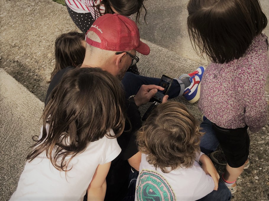 Un homme avec une casquette entouré d'enfants qui regardent lénigme d'une chasse au trésor géante en réalité augmentée sur son téléphone